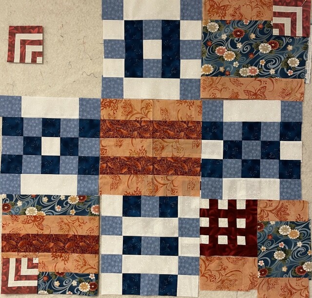 In this one, the blue, dark blue, and white squares are still top and bottom and central side spots. 

In the middle is the light orange and dark orange batik striped square. 

On the top right is the light orange batik with blue floral Japanese print in the middle. Now there is a corner square of alternating white and red lines in a chevron pattern. 

Bottom right has a grid of red and white at the top left corner of the block and then there is light orange, blue floral Japanese print, and more light orange batik. 

Lower left has blue floral Japanese print on the outside, two strips of light orange batik, and then dark orange batik in the middle. At the lower left is another square made of alternating red and white strips in a chevron pattern.