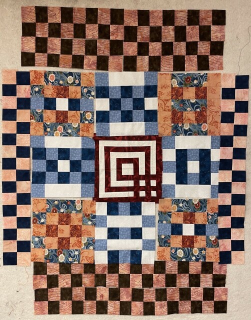 Checkerboard quilt made with alternating pink, blues, whites, and browns. There’s a more complex maze like square in the middle made with red and blue. 