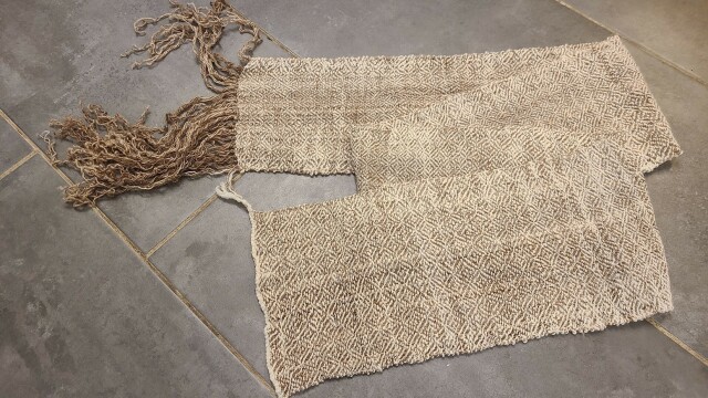 A length of brown and cream fabric about the size of a scarf.  It has an all over diamond pattern, a fringe on one end, and a woven hem-band on the other