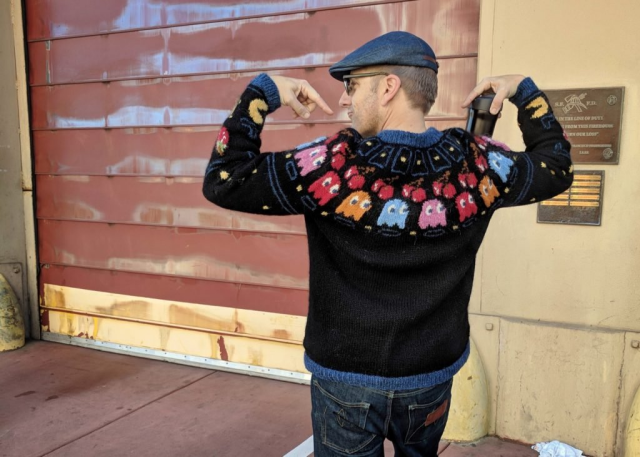 Me showing off my Pac-Man sweater. It is a hand-knit sweater in an Icelandic lopapeysa style. Most of the sweater is black, but near the neck is a row of ghosts, cherries, and other Pac-Man iconography. 