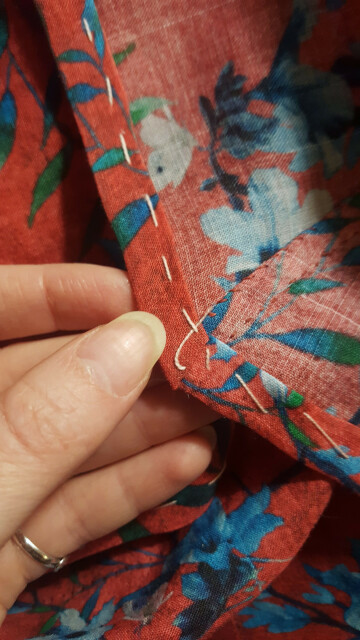 A very nicely mitred hem basted down with loose light pink basting stitches. There is a thicker bit of seam at the corner, and the different layers have been fussed with until a very neat and well behaved looking intersection of all the material has been achieved.