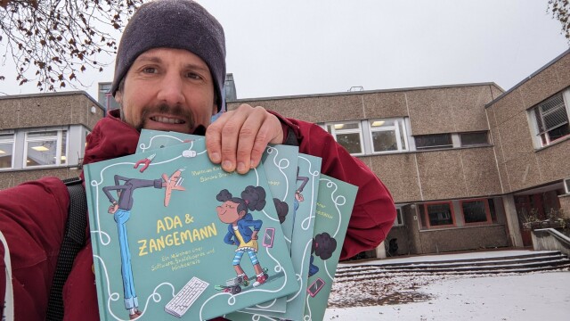 Person holding four books "Ada&Zangemann" in German, Arabic, Ukrainian, and Italian in front of school building with the ground covered with snow.