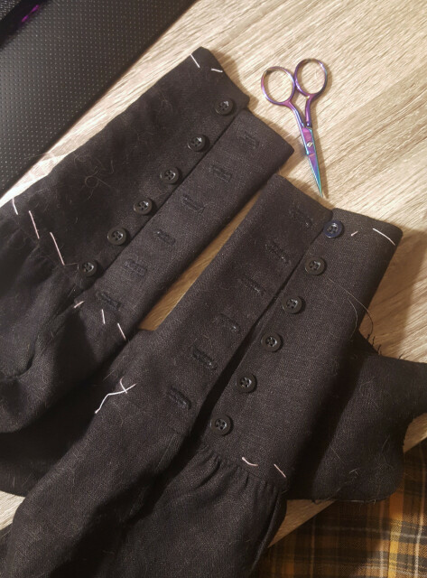 A photo of two sleeves with their cuffs laid out on a table with pale wood grain. The sleeves have been neatly gathered to the cuffs, which are very tall and narrow, and will envelop most of the forearm when worn. They have twelve buttonholes and buttons between them. The rows of them look nice and neat. The buttonholes look pretty uniform but not perfectly, indicating more organic hand sewing. The buttons are a plain black with a wide rim. There is pink basting thread along the top and wrist edge of the cuffs.