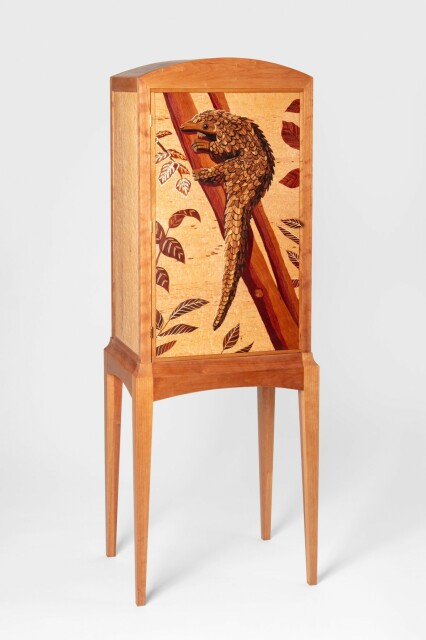 Photograph of a tall wooden cabinet. The door of the cabinet has a marquetry image of a pangolin climbing a tree. The cabinet has long legs and stands about six feet tall.