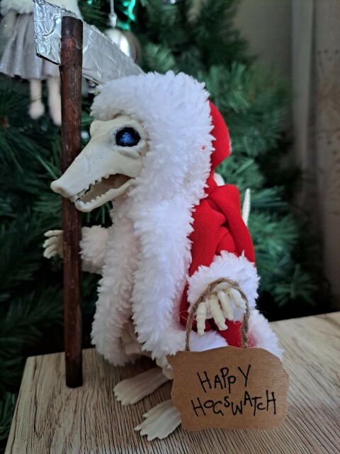 The left hand view of a plastic skeleton rat standing upright on its back legs. The rat is wearing a red and white cape as seen on Santa. In its left hand is a sign saying Happy Hogswatch. In its right hand can be seen the handle and top of the blade of a scythe.