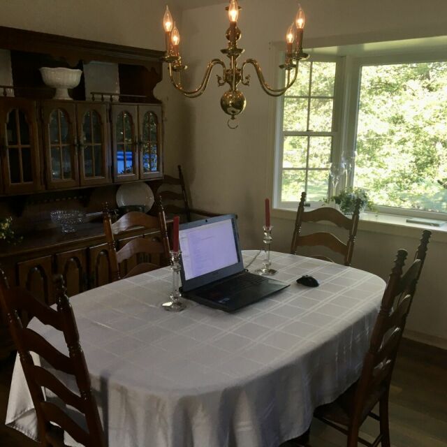 A laptop computer on a dining table covered with a white linen tablecloth and flanked by a couple of glass candle holders. In the background, there’s a wooden hutch on the left, and a large bay window looking out on a lush tree on the right. Above the table hangs a brass chandelier with lit faux-candles.