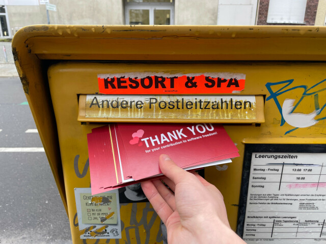 Photo of my hand while inserting many red postcards in a yellow post box in Berlin. The postcards have a writing that says “THANK YOU for your contribution to software freedom!”