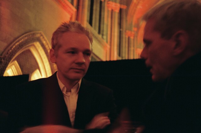 An analog photo taken in 2011 of Julian Assange whispering to the hear of Kristinn Hrafnsson. The background is the gothic, cathedral-esque High Court of Justice in London. The colour are yellow/red denoting an antique lighting. 

The photo is CC BySa (or LAL 1.3) J. Zimmermann