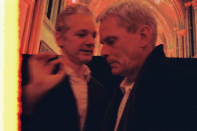 An analog photo taken in 2011 of Julian Assange whispering to the hear of Kristinn Hrafnsson. The background is the gothic, cathedral-esque High Court of Justice in London. The colour are yellow/red denoting an antique lighting. The edge of the frame is burnt, indicating a recently loaded film, and blur on Assange's hand and parts of his face evokes a rather dynamic conversation.

The photo is CC BySa (or LAL 1.3) J. Zimmermann