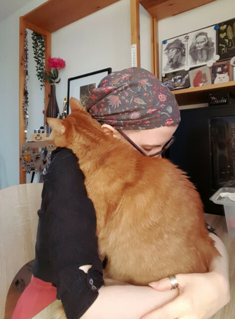 A slightly low quality but quite colourful and cute photo of a ginger tabby cat sitting between the forearms of Sini. He has his chin and head resting on her shoulder, leaning against her chest. She has her head down and cheek leaning against his flank. The photo is a bit blurry but his fur is a blazing warm copper tone, and there is pleasant colour harmonies of the red details in her black, red and grey clothes, and the decorations and postcards in the background.