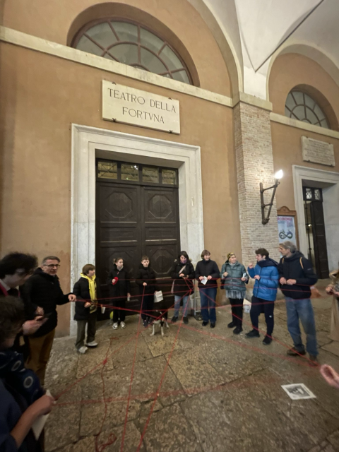 Persone in cerchio di fronte al Teatro della Fortuna di Fano, mentre tengono capi di un gomitolo rosso che le connette.