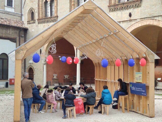 A wider picture of the reading, in a wooden structure with FSFE red and blue balloons hanging, and people of various ages listening.