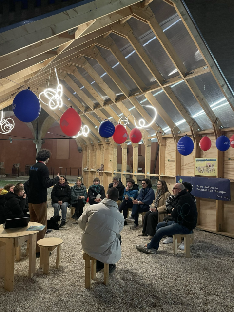 A wooden house by night, lit by special white lights. FSFE balloons hanging, a group of people seated in circle, and Tommi standing while talking.