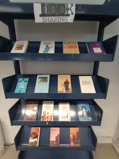 Lo scaffale del booksharing in biblioteca, tra gli altri libri di Deaglio, Baricco, Bulgakov, Benni, Maurensig