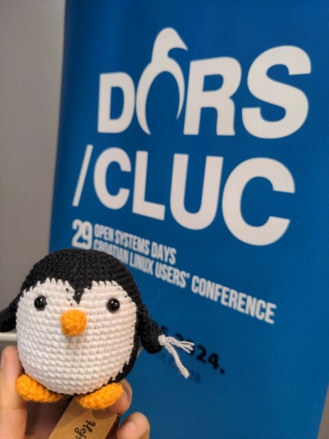 Image showing a crochet Linux penguin with the background of the DORS/CLUC conference banner 