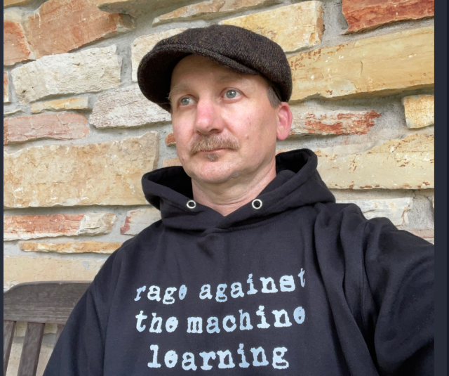 man with cap and black sweatshirt with gray letters saying "Rage Against the Machine Learning."