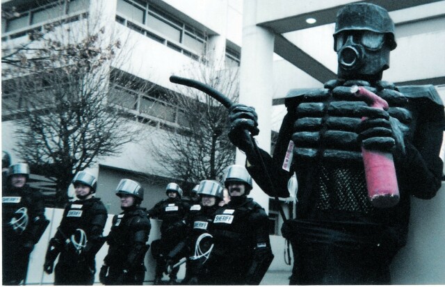 Line of riot cops in front of the Seattle Jail at the WTO anniversary in 2000. Standing next to a 12 foot tall riot cop puppet. One cop with a moustache is smiling for the camera.