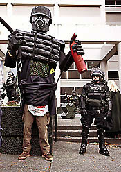 Another photo of Officer Friendly, the giant riot cop standing next to his mascot, the much shorter real riot cop in front of the Seattle jail. This image shows Officer Friendly’s puppeteer legs looking rather uninspiring in unmatched brown work pants. Also the carpenters nail bag wrapped around the puppeteer’s waist, which held the super soaker water jug that supplied the working “pepper spray” gun.