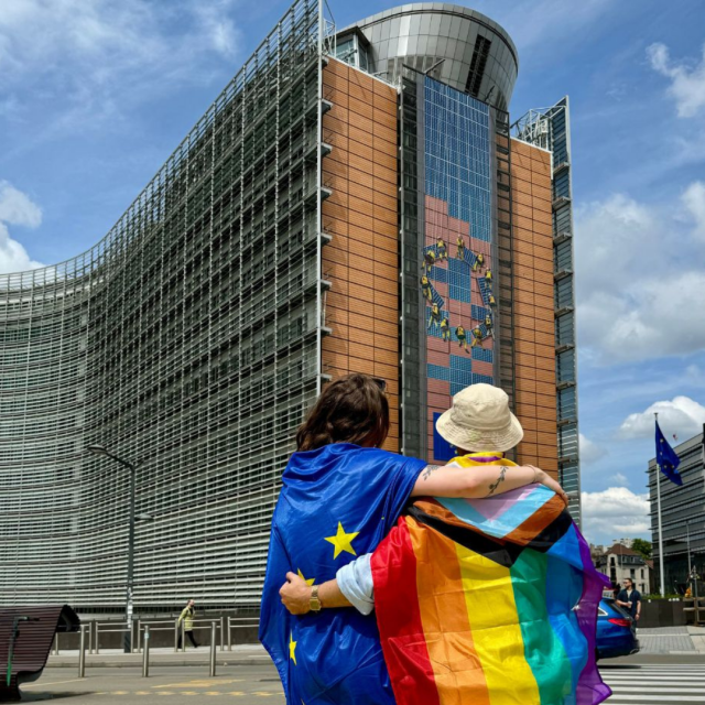 A photo of two people, one draped in the EU flag and the other draped in the rainbow flag. They are both standing in front of the European Commission headquarters, the Berlaymont, in Brussels. 

