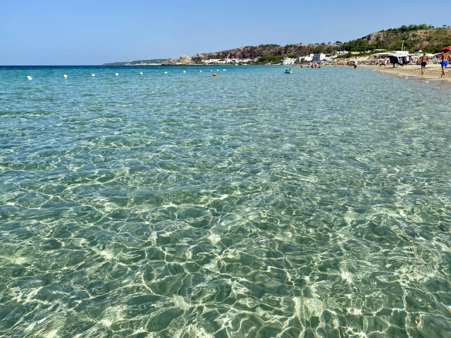 
A clear beach scene with transparent turquoise water in the foreground, showcasing the sun's reflections on the water's surface. The beach is lined with people enjoying the day, with some under umbrellas and others walking along the shore. The background includes rocky hills and a few buildings, all under a clear blue sky.