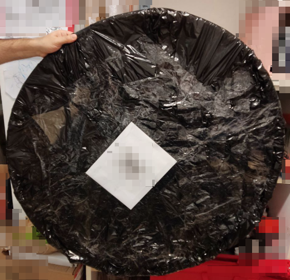 foto di un pacco a forma di cerchio imballato con plastica nera, diversi strati di nastro adesivo trasparente e foglio A4 bianco attaccato con l'etichetta del corriere. Il "cerchio" è tenuto sollevato dalla mano di Yaku