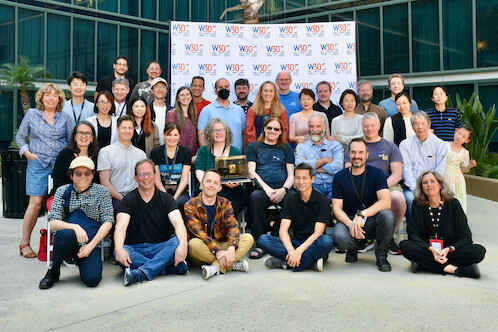 Group photo of the W3C Team: 36 people of mixed gender, age and race posing in front of a white backdrop with repeated W30C logos amid palm trees in a courtyard