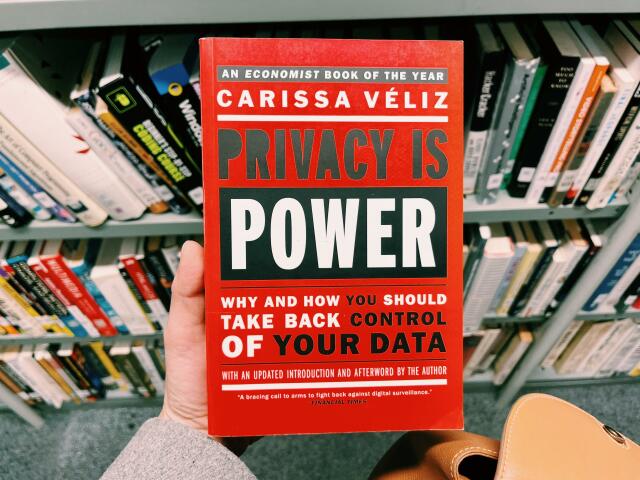 A photo of a hand holding up the book “Privacy is Power. Why and how you should take back control of your data” by Carissa Véliz. You can see rows of books in the background 
