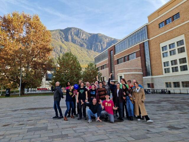 Group of people assembled for group picture, doing some fun staff in front of building and mountain landscape. 