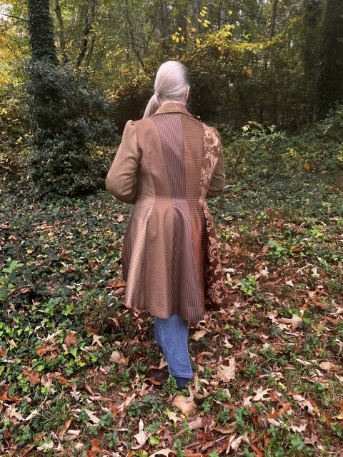 Me standing outside in autumn leaf covered grass, with my back to the camera, wearing a brown coat with a hemline that's shorter in the front than the back. The coat has vertical panels that are made of different fabrics, some print and some solid, all in different shades of brown.