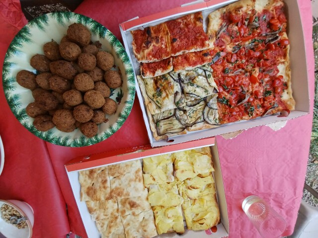 Due cartoni di pizza al taglio vari gusti, una scodella di falafel, un bicchiere di vino bianco. Tovaglia rossa. Questa inquadratura si chiama plongée