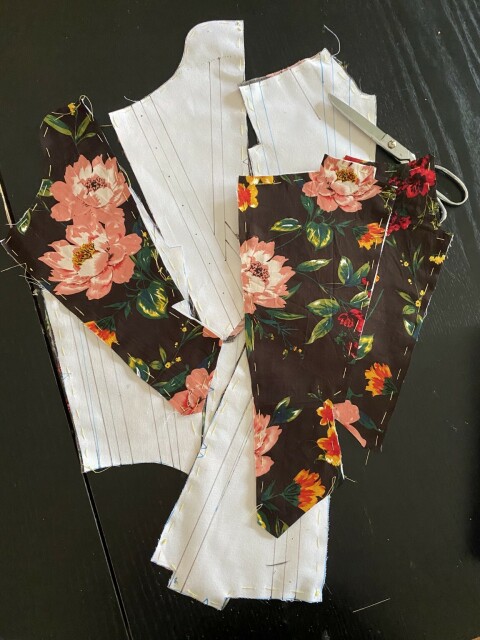 Pieces cut out for 18th century stays. The outer fabric has a bold floral pattern with big pink peonies and smaller sprays of red, orange, and yellow flowers on a dark brown background. The interlining is white coutil, marked with lines for boning channels and eyelets. The pieces are basted together loosely with yellow thread.