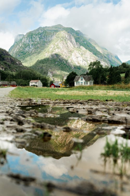 A ground level shot across a puddle of water looking towards some houses at the end of a road, with a rocky mountain rising behind. The mountain is reflected in the water.