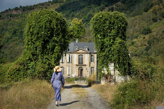 An overgrown gravel track leading towards a two story house that's nestled between some trees, with a hillside beyond. There's a person in a long blue dress and straw hat walking away from the camera towards the house.