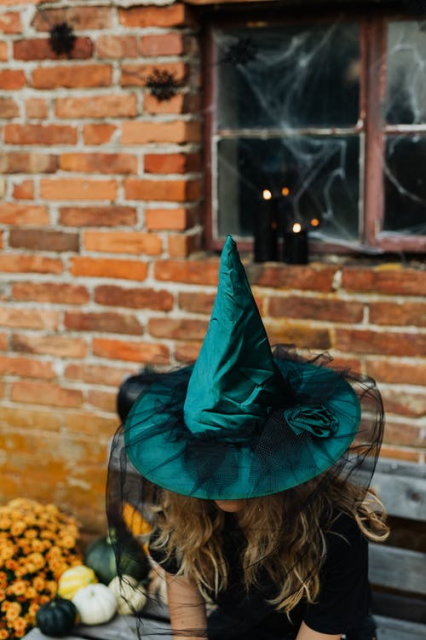 A person in a dark green pointy witch hat with a black veil sitting on a wooden bench next to an assortment of pumpkins, with a red brick wall and an old wooden window frame covered in cobwebs behind. The wall has some halloween decorations. The person has long blonde hair that's just visible under the wide brim of the hat, and they're wearing a black top. 