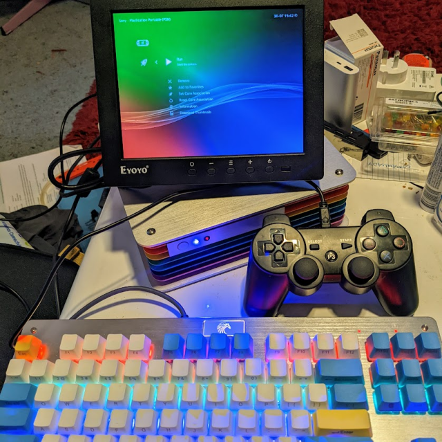 A lit up keyboard with RGB LEDs in it attached to an aluminium PC case with rainbow coloured fins around the edge, and a monitor showing a menu with an RGB background pattern