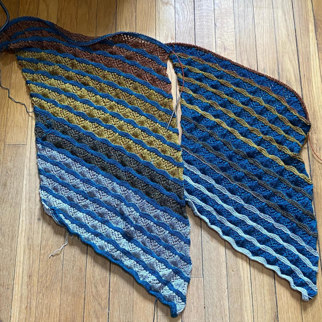 Two lace shawls in progress, in alternating colors of a gray-to-yellow-to-brown gradient and a turquoise color. They are alternating sections of lace and garter stitch, and the color placements are swapped between the two shawls.