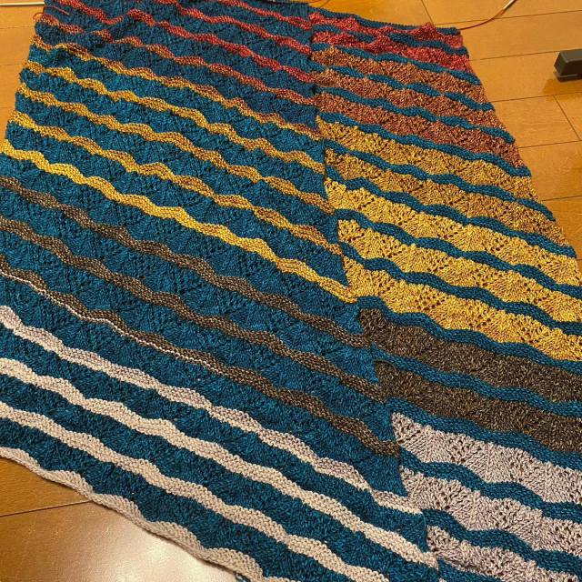 Two knit lace shawls in progress, alternating between sections of lace and garter stitch. The colors alternate between a band of turquoise and a gradient moving from gray to yellow to red, and the placement of the bands is swapped between the two shawls.